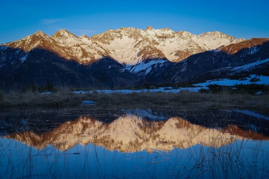 Snow-covered peaks rise majestically against a clear blue sky, with their reflection captured in still water at dawn in a peaceful alpine setting.