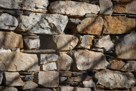 A solid stone wall displays a variety of rock shapes and colors, illuminated by sunlight filtering through trees. Shadows create an interesting pattern on the surface.