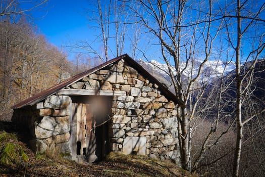 A rustic stone cabin sits against a backdrop of majestic mountains, with skeletal trees surrounding it under a crisp blue sky. The setting conveys tranquility and solitude in nature.