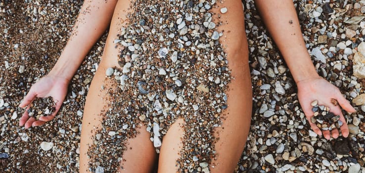 A nude woman reclines on a sandy beach, her body partially covered with small pebbles. She is relaxed, enjoying the warmth of the sun on a clear day.