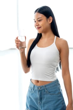 Vertical image of beautiful Asian woman hold glass of water and stand in front of glass window with day light and she look happy with look at the glass.