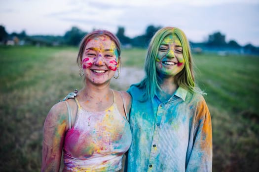 Two vibrant young women, coated in colorful powder, embrace and share smiles at the holi festival festivities