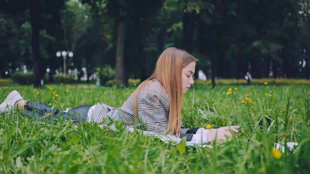 Young student working on her laptop while lying on the grass in a park, enjoying the fresh air and green environment