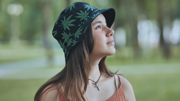 Portrait of a teenage girl wearing a bucket hat with cannabis leaf pattern, looking up in contemplation in a summer park