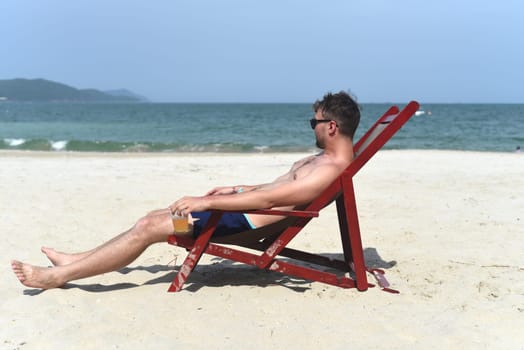Young man drinking beer and sitting on deck chair at beach in Nha Trang