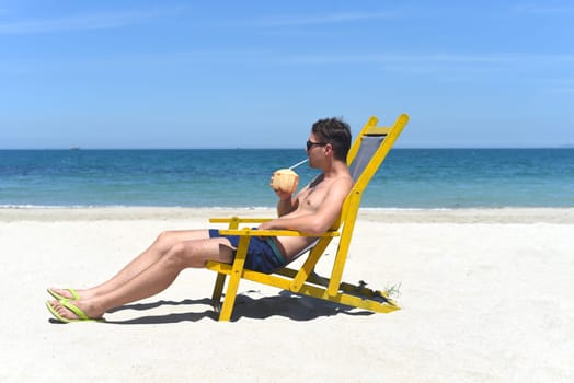 Young happy man drinks coconut juice sitting on deck chair on a south china sea beach in vietnam
