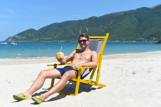 Young happy man drinks coconut juice sitting on deck chair on a south china sea beach in vietnam