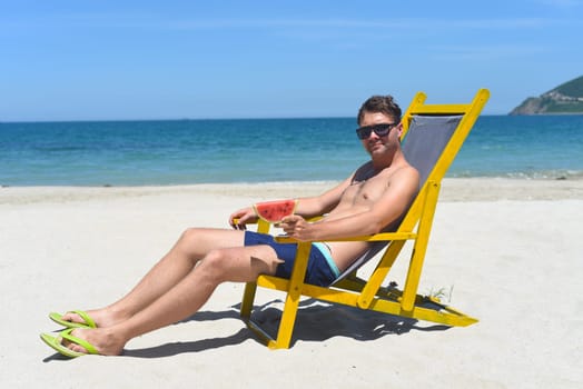 Young happy man eating watermelon sitting on deck chair on a south china sea beach in vietnam