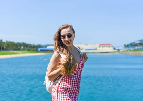 Bright blond young woman standing with backpack near sea in summer