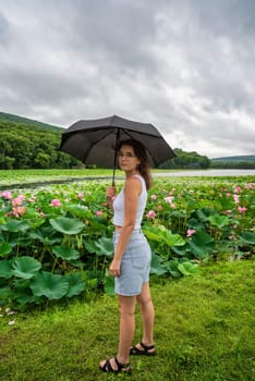 Upset woman holding blak umbrella under rain and enjoying rainy weather near lotus lake in the Far East of Russia
