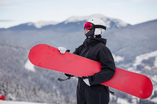A snowboarder in black gear holding a red snowboard on a snowy mountain with a scenic view.