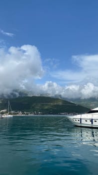 A tranquil harbor with yachts docked, clear reflections on the water, and mountains under a bright blue sky with fluffy clouds.