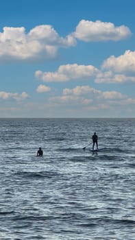 Silhouette of two people on a stand up paddle board in calm open water under a bright blue sky with fluffy clouds.