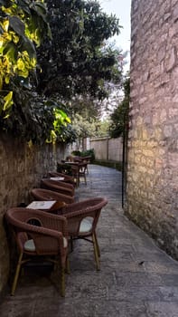 A narrow cobblestone alley with wicker chairs and tables set against stone walls, surrounded by greenery. . High quality photo