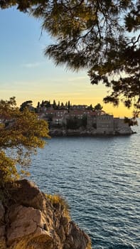 Sveti Stefan, a small island and resort in Montenegro.Stone buildings and lush greenery, view from a rocky shore framed by pine trees at sunset.
