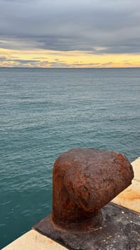 A rusted bollard on a stone pier overlooking calm blue sea waters under a cloudy sunset sky, with golden hues on the horizon.