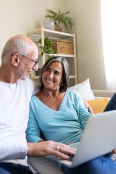 Vertical portrait of mature married couple in love using laptop relaxing on the sofa looking at each other smiling. Lifestyle and technology concepts.