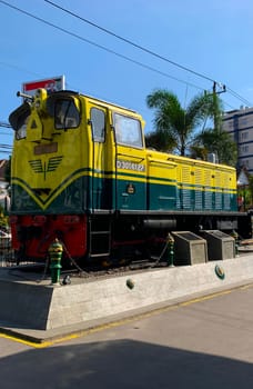 Bright yellow and green antique Diesel locomotive D30162 on display used to be maintained by Kereta Api Indonesia, showcasing railway history in a city setting. Features urban surroundings, clear blue skies, and represents both transportation innovation and nostalgia for vintage trains.