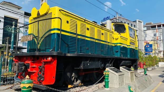 Bright yellow and green antique Diesel locomotive D30162 on display used to be maintained by Kereta Api Indonesia, showcasing railway history in a city setting. Features urban surroundings, clear blue skies, and represents both transportation innovation and nostalgia for vintage trains.