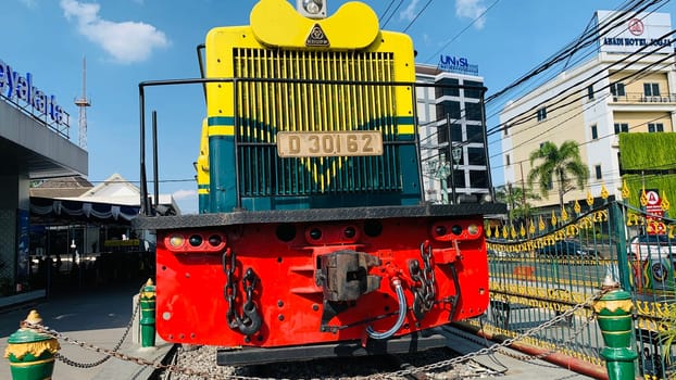 Bright yellow and green antique Diesel locomotive D30162 on display used to be maintained by Kereta Api Indonesia, showcasing railway history in a city setting. Features urban surroundings, clear blue skies, and represents both transportation innovation and nostalgia for vintage trains.