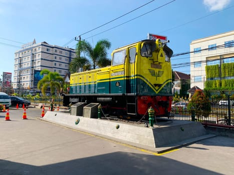 Bright yellow and green antique Diesel locomotive D30162 on display used to be maintained by Kereta Api Indonesia, showcasing railway history in a city setting. Features urban surroundings, clear blue skies, and represents both transportation innovation and nostalgia for vintage trains.