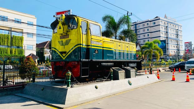 Bright yellow and green antique Diesel locomotive D30162 on display used to be maintained by Kereta Api Indonesia, showcasing railway history in a city setting. Features urban surroundings, clear blue skies, and represents both transportation innovation and nostalgia for vintage trains.
