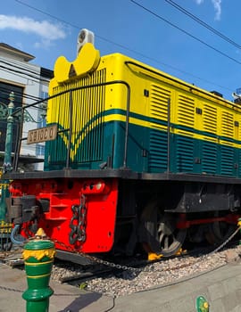 Bright yellow and green antique Diesel locomotive D30162 on display used to be maintained by Kereta Api Indonesia, showcasing railway history in a city setting. Features urban surroundings, clear blue skies, and represents both transportation innovation and nostalgia for vintage trains.