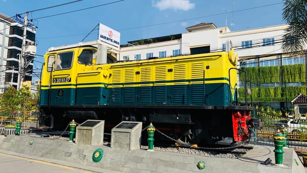 Bright yellow and green antique Diesel locomotive D30162 on display used to be maintained by Kereta Api Indonesia, showcasing railway history in a city setting. Features urban surroundings, clear blue skies, and represents both transportation innovation and nostalgia for vintage trains.