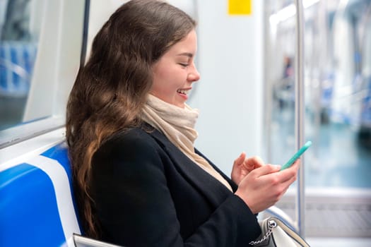 Cheerful young woman in a black coat and beige scarf sitting inside a modern subway train, laughing while looking at her smartphone. She enjoys a fun moment during her commute.