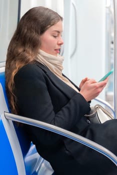 Cheerful young woman in a black coat and beige scarf sitting inside a modern subway train, laughing while looking at her smartphone. She enjoys a fun moment during her commute. Vertical