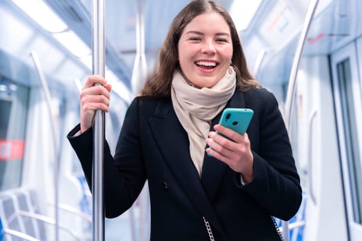 Young woman in a black coat and beige scarf standing inside a modern subway train, holding a smartphone and gripping a metal pole. She appears focused while commuting, carrying a stylish white purse.