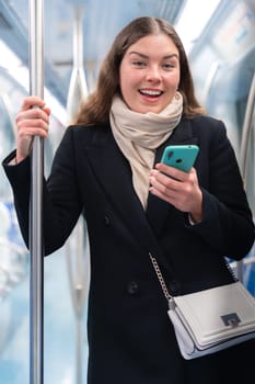 Cheerful young woman in a black coat and beige scarf talking on her smartphone while standing inside a modern subway train. She smiles brightly, holding onto a metal pole during her commute.