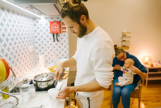 A man expertly cooks in a modern and stylish kitchen while a woman lovingly holds a baby, showcasing family life
