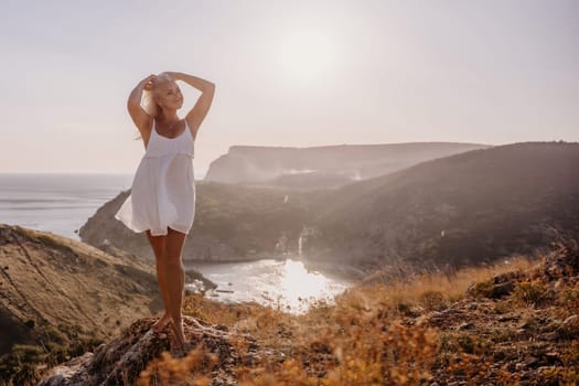 A blonde woman stands on a hill overlooking the ocean. She is wearing a white dress and she is enjoying the view