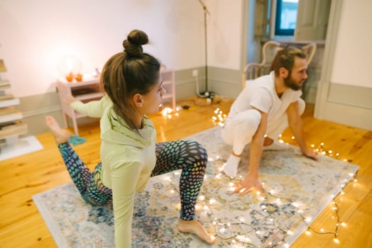 Two individuals are engaging in yoga poses within a cozy and warmly lit room adorned with enchanting fairy lights
