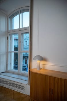 A room with a large window. bright room with a large window and a lamp on the chest of drawers. the lamp glows with a warm light. the chest of drawers is made of natural wood in a pleasant beige shade
