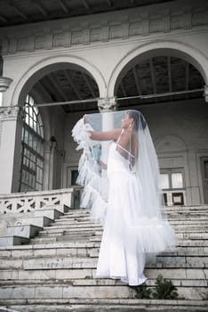 beautiful bride with a bouquet of flowers covered with a veil. a beautiful girl stands on the steps of a light building. she stands sideways in a white long dress and lifts the veil with her hands