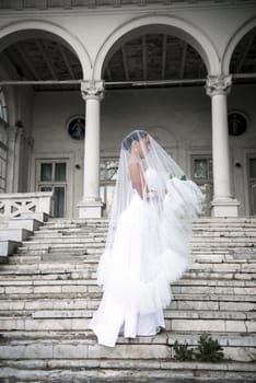 beautiful bride with a bouquet of flowers covered with a veil. a beautiful girl stands on the steps of a light building. she stands sideways in a white long dress.full length general plan