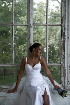 A beautiful bride in a long elegant dress with a bouquet of lilies of the valley sits near the window. A brunette girl with a beautiful hairdo, she smiles. She has white flowers and a white dress.