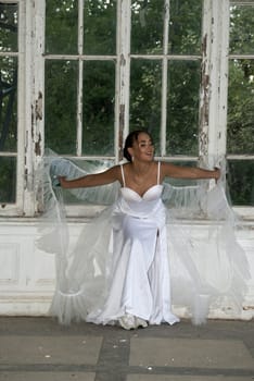The bride is sitting near the window, she has a lush veil and an elegant dress. The girl is a brunette with a smooth hairstyle. She lifts the veil with her hands and smiles.
