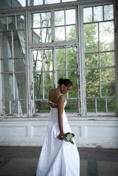 A beautiful bride in a long elegant dress with a bouquet of lilies of the valley stands near the window. A brunette girl with a beautiful hairdo, she turned around. She has white flowers