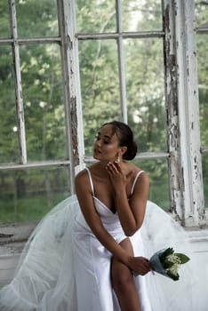 beautiful bride in a long elegant dress with a bouquet of lilies of the valley sits near the window. brunette girl with beautiful hair styling. she has a lush elegant dress with straps