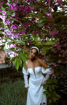very beautiful girl among blooming sakura. girl with beautiful make-up and hairstyle looks at camera. girl is wearing long white dress she has dark eyes. pink flowers around her