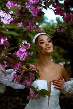 beautiful girl in white dress among pink flowers. she has stylish hairstyle with white elements, dress with open shoulders with smooth tanned skin. she smiles, she has beautiful makeup