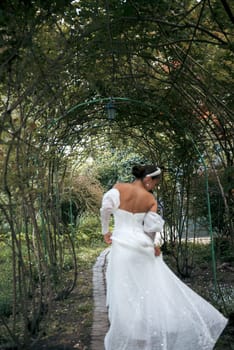 very beautiful girl in a long white dress, with a stylish hairstyle with white decorations in her hair. against the background of an arch of greenery. she leaves, view from behind