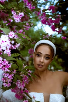 beautiful girl in white dress among pink flowers. she has stylish hairstyle with white elements, dress with open shoulders with smooth tanned skin. girl looks into the frame she has beautiful makeup