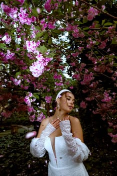 beautiful girl in white dress among pink flowers. she has stylish smooth hairstyle with white elements she is brunette. dress has open shoulders with smooth tanned skin. pink sakura petals fall on her