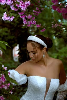 Beautiful girl in white dress among pink flowers. She has stylish hairstyle with white elements, dress with open shoulders with smooth tanned skin. Waist is emphasized by corset.