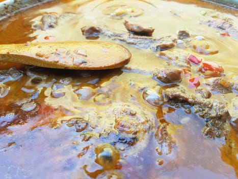 Close-up of bubbling beef stew with meat, vegetables, and rich broth in a pot. A wooden spoon rests in the liquid, showing the simmering cooking process. Rustic outdoor setting highlights traditional comfort food.