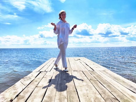 Mature adult female blogger walks on a wooden dock by the water, embracing nature on her journey under a bright sky.